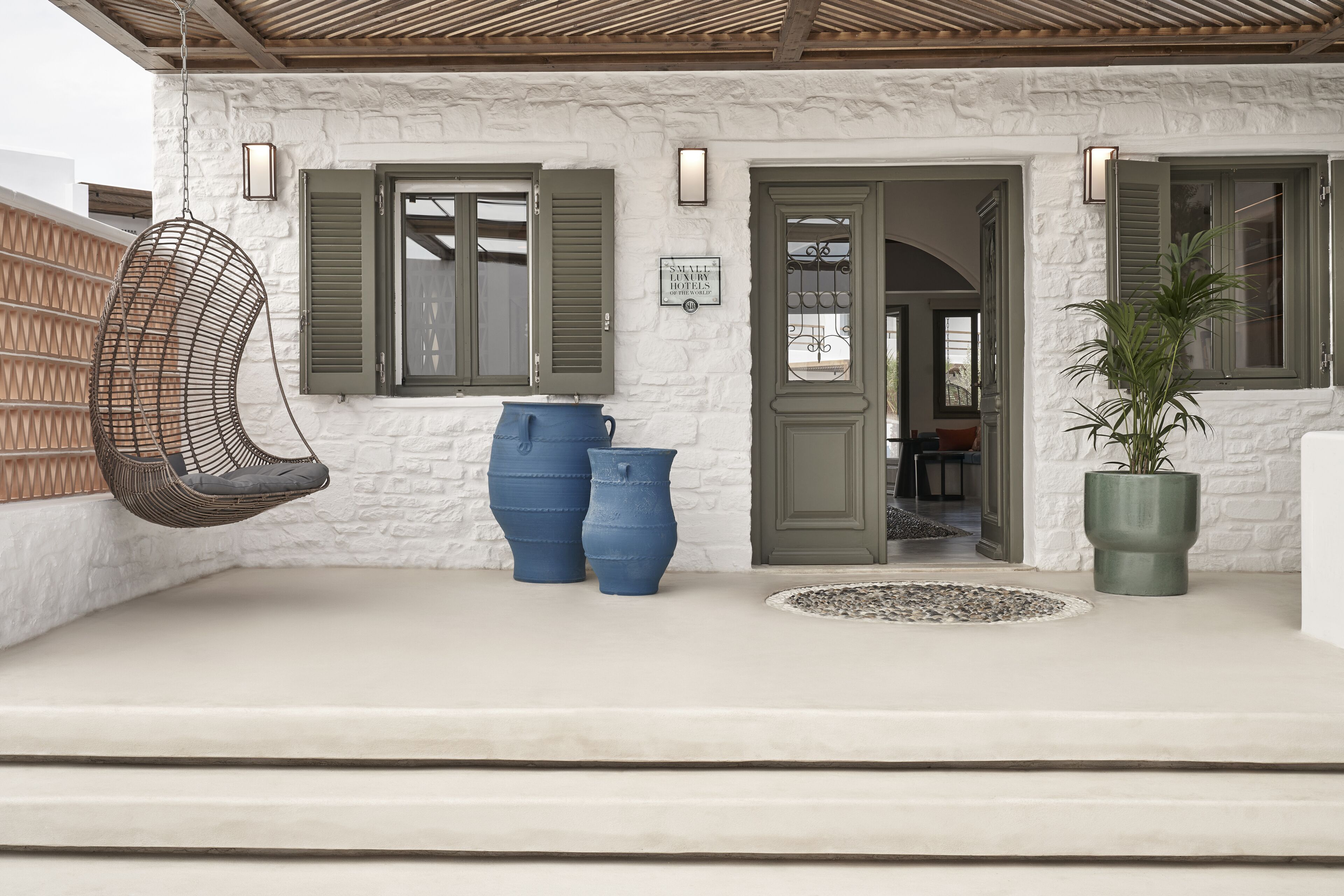 A white building with green shutters, a hanging chair, two blue planters, and a potted plant.