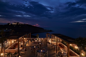 An outdoor dining area with tables and chairs, surrounded by wooden structures and plants. - Andronis Minois (Paros)