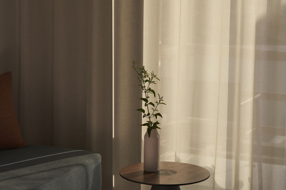 Una sala de estar moderna con un sofá, una mesita redonda con un florero y libros, y cortinas translúcidas que dejan entrar luz natural.