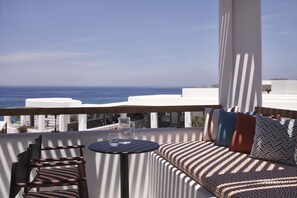 Un balcon avec vue sur la mer, une table ronde avec deux verres d’eau et un canapé agrémenté de coussins à motifs.