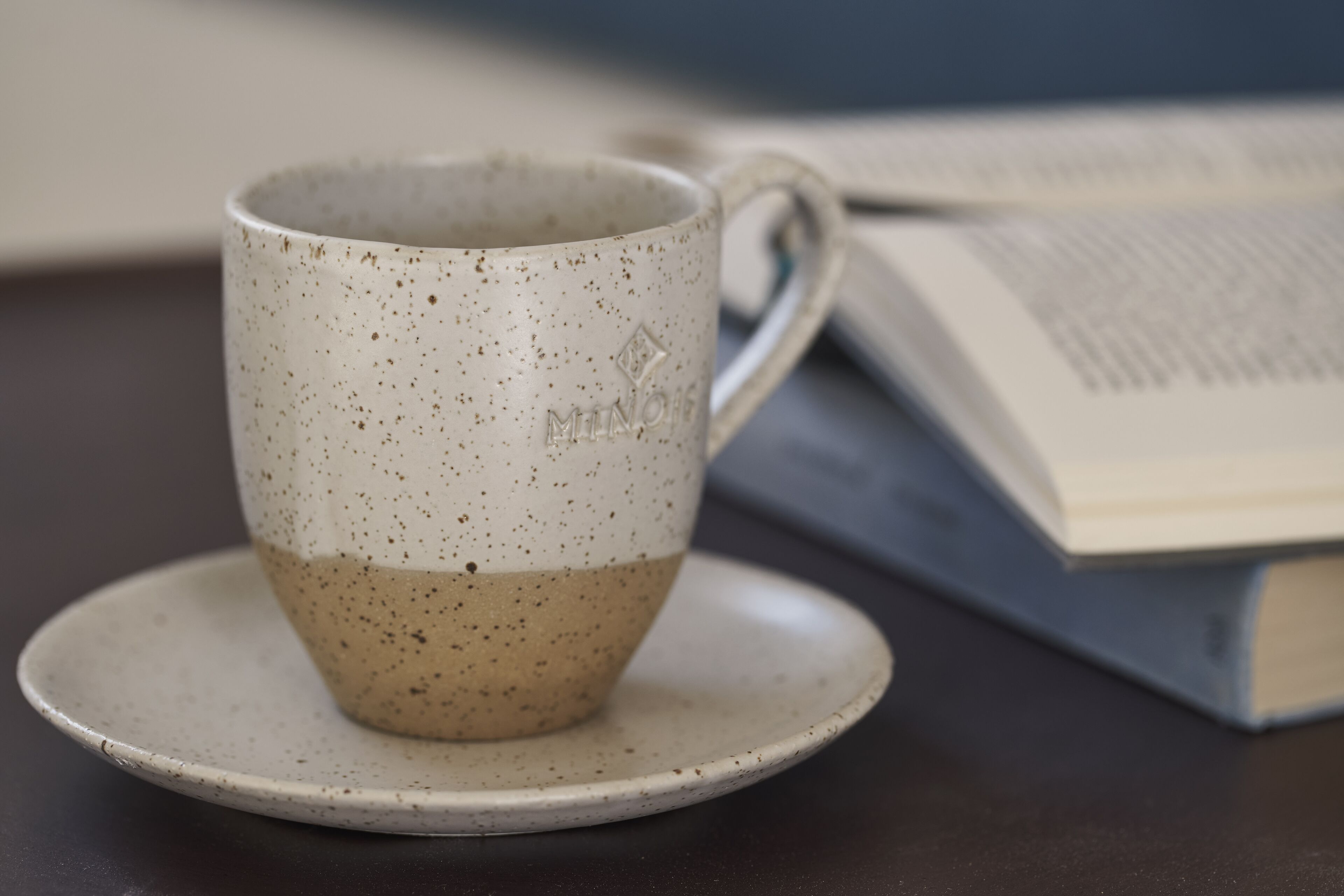 A ceramic mug with a speckled design on a saucer, placed on a table with a book nearby.