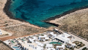 An aerial view of a coastal resort with white buildings, a swimming pool, and clear blue waters.