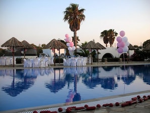 A pool area with white tables and chairs, thatched umbrellas, and pink and white balloons.