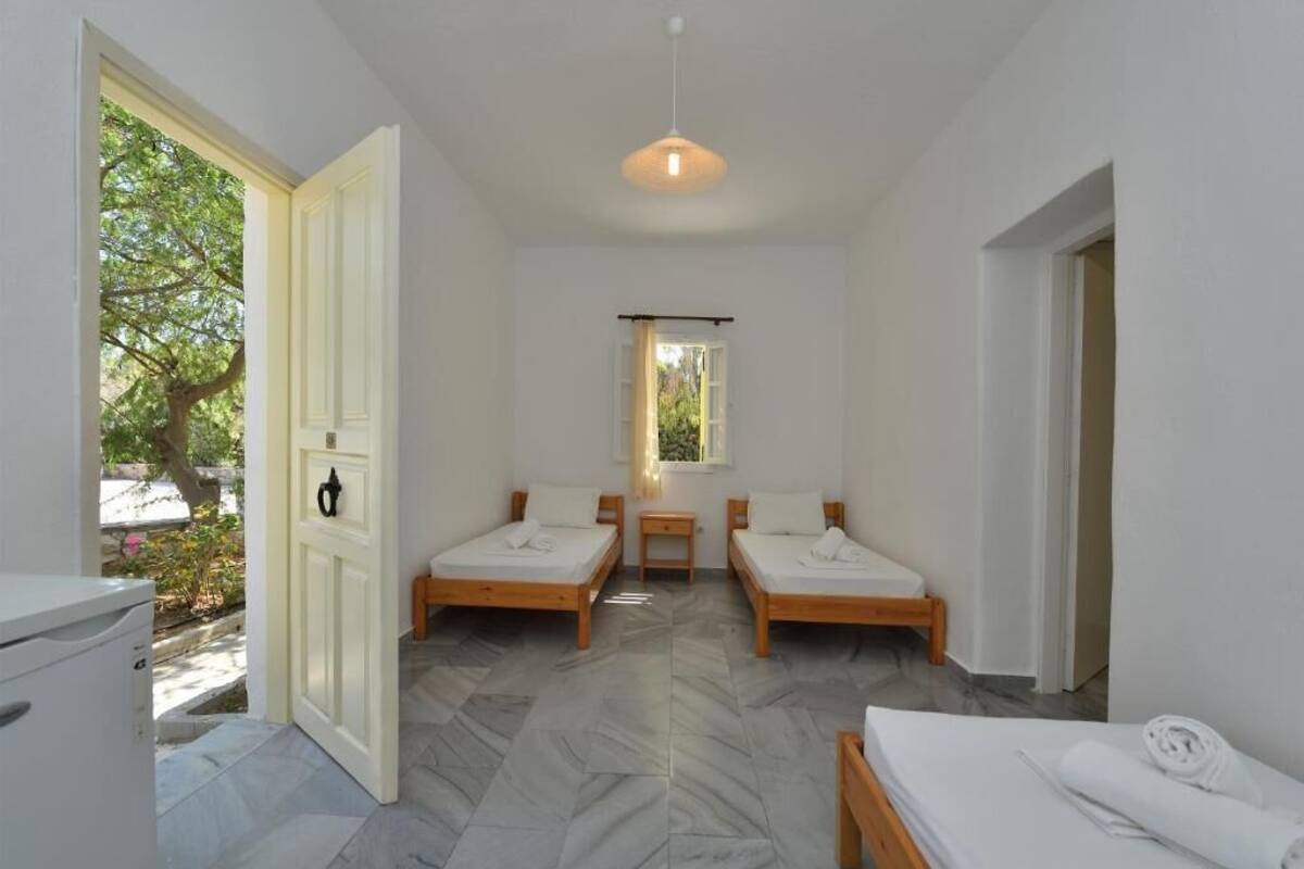 Ein Zimmer mit zwei Holzbetten, einem kleinen Tisch, einem Fenster mit Vorhängen und einer zur Außenwelt führenden Tür.