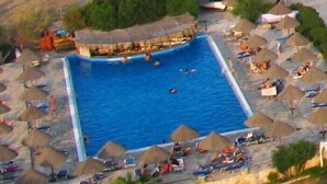 An aerial view of a square swimming pool surrounded by beach umbrellas and lounge chairs, with a building adjacent to the pool.