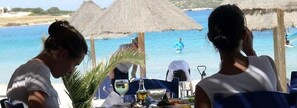 Un área de comedor frente a la playa, con sombrillas de paja, una mesa dispuesta con vasos y una botella, y vista al mar.