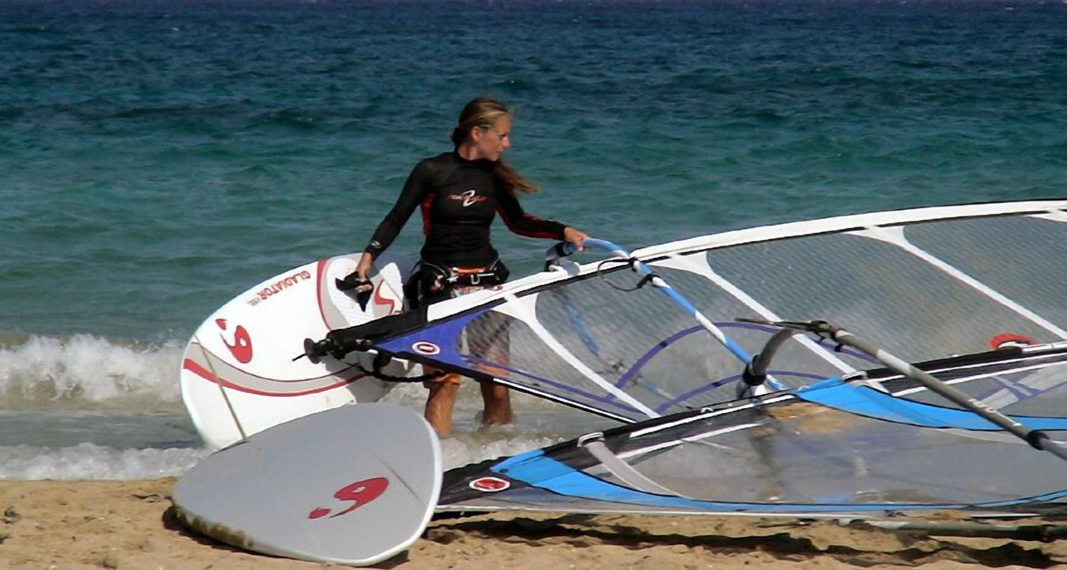 Una persona porta una taula de windsurf i una vela a la platja.