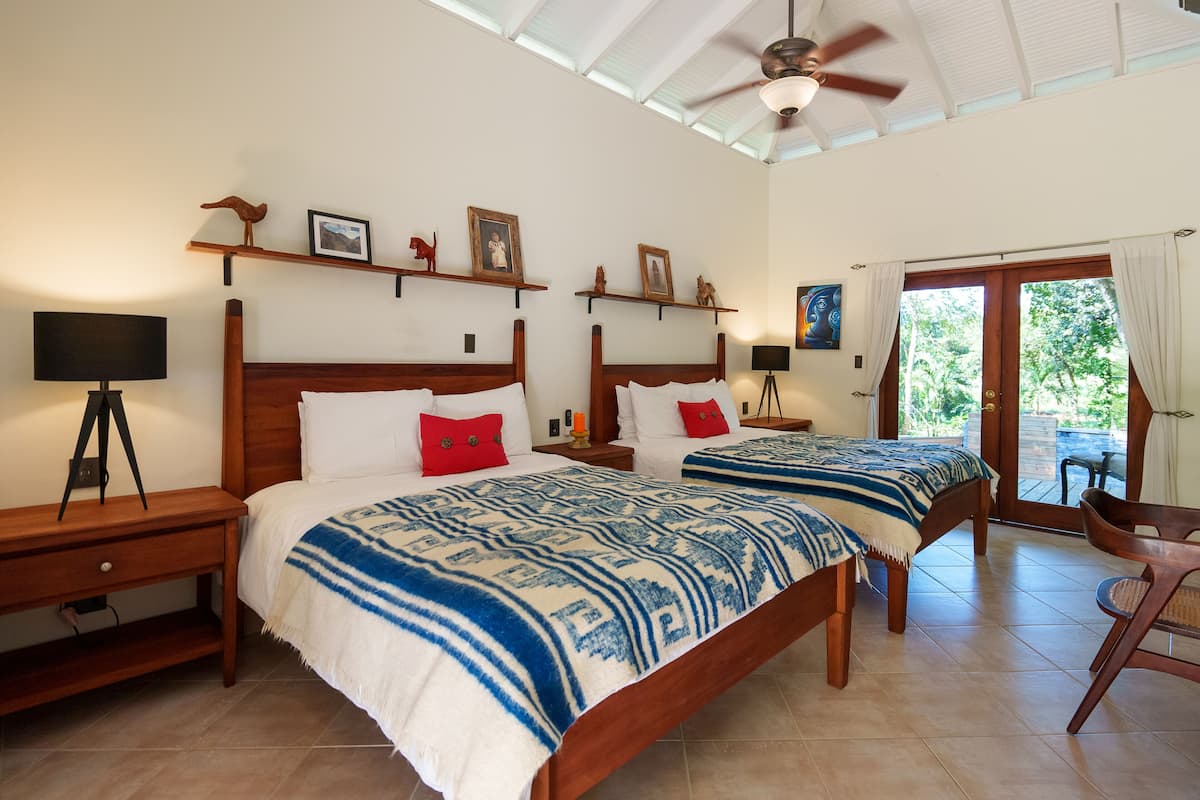 Habitación doble con dos camas, cabeceros de madera y ventilador de techo. Hay repisas con objetos decorativos y vista al exterior a través de una puerta de vidrio.
