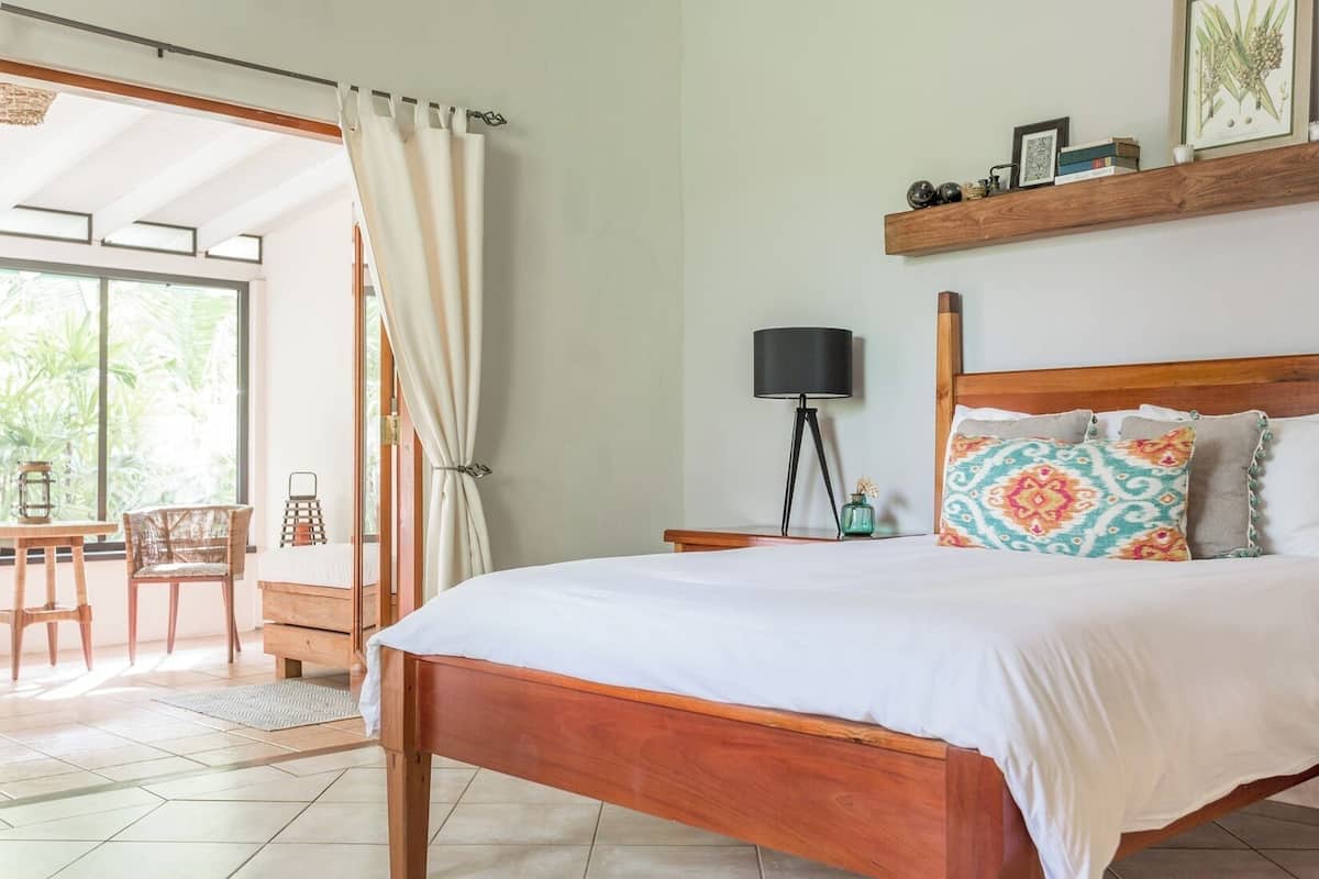 Un dormitorio con una cama de madera, una mesita de noche con una lámpara, una silla y vistas al exterior.