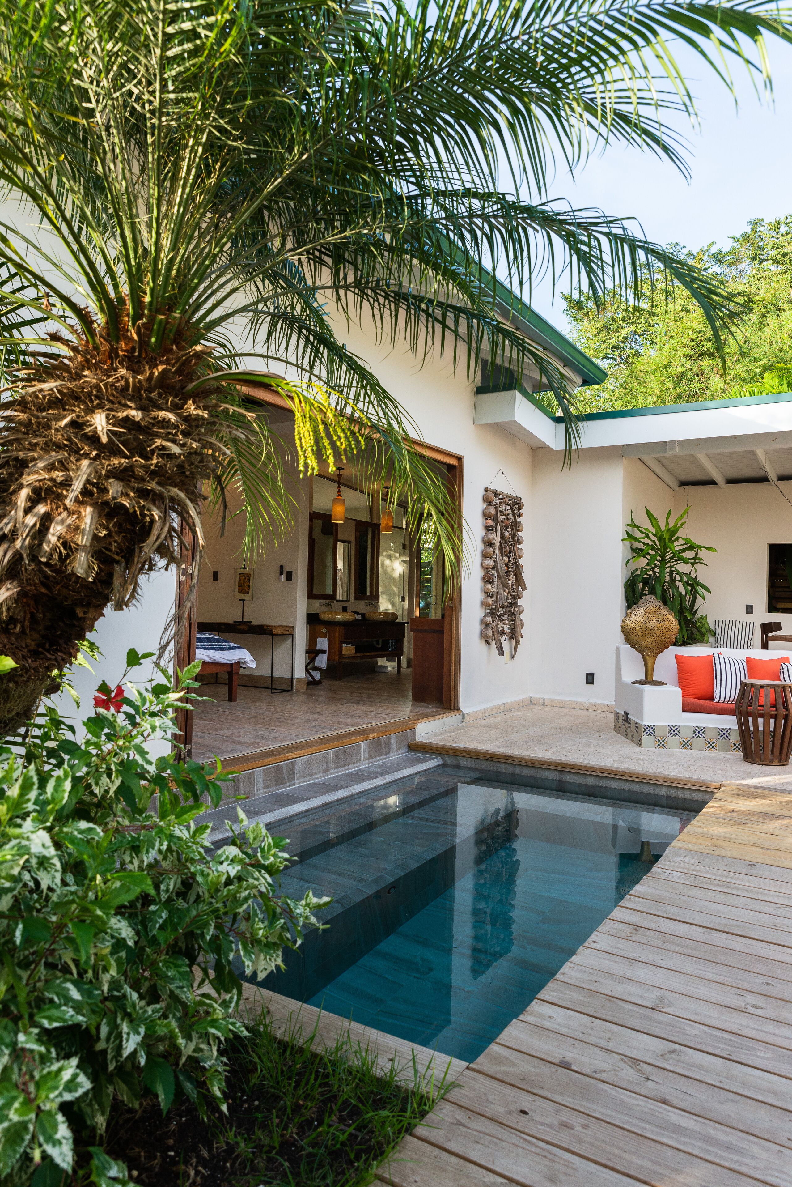 A modern outdoor pool area with a wooden deck, a palm tree, and a cozy seating area with a sofa and chairs.