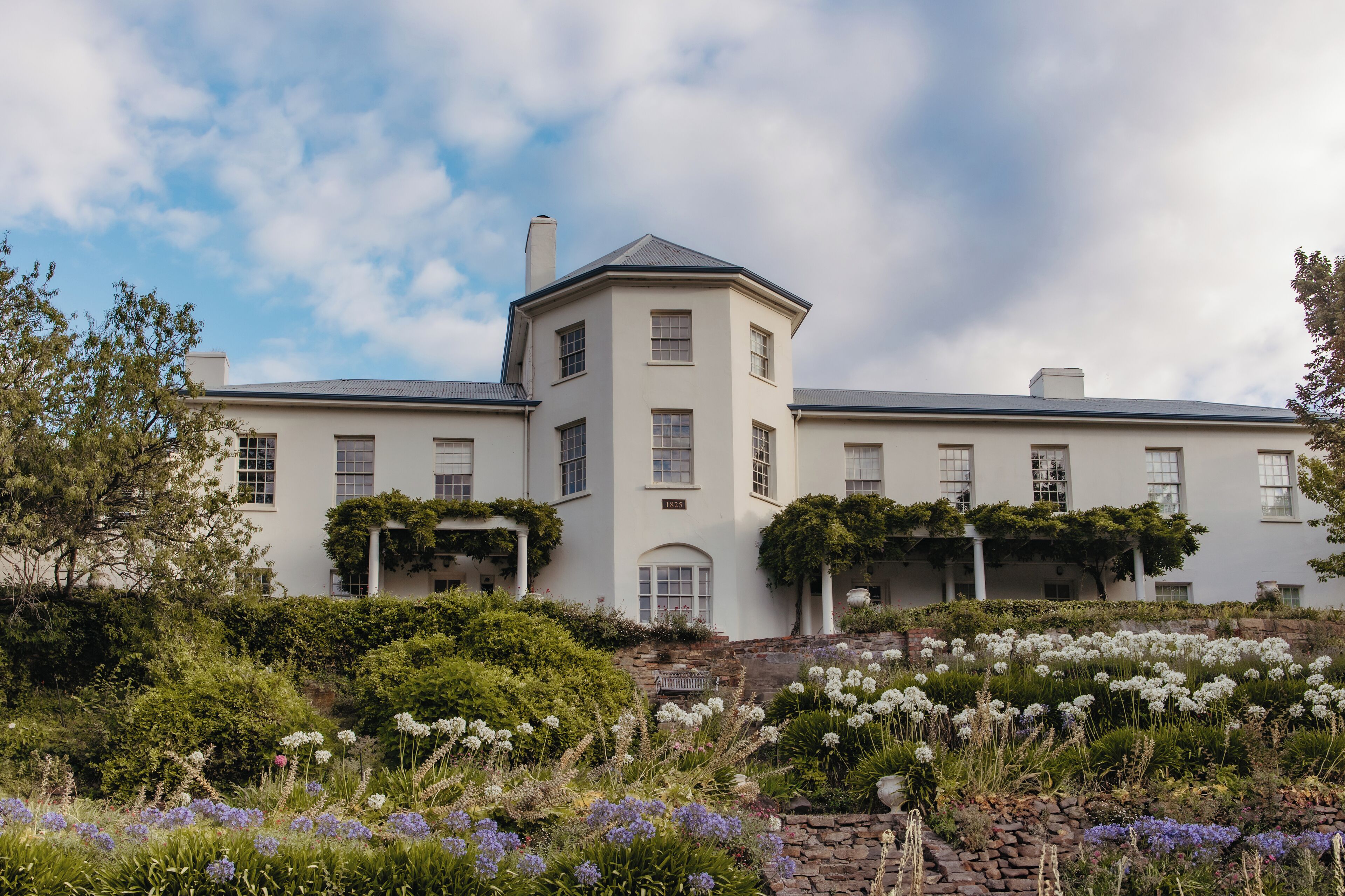 A large, white, multi-story building with a central tower, surrounded by lush gardens and flowering plants.