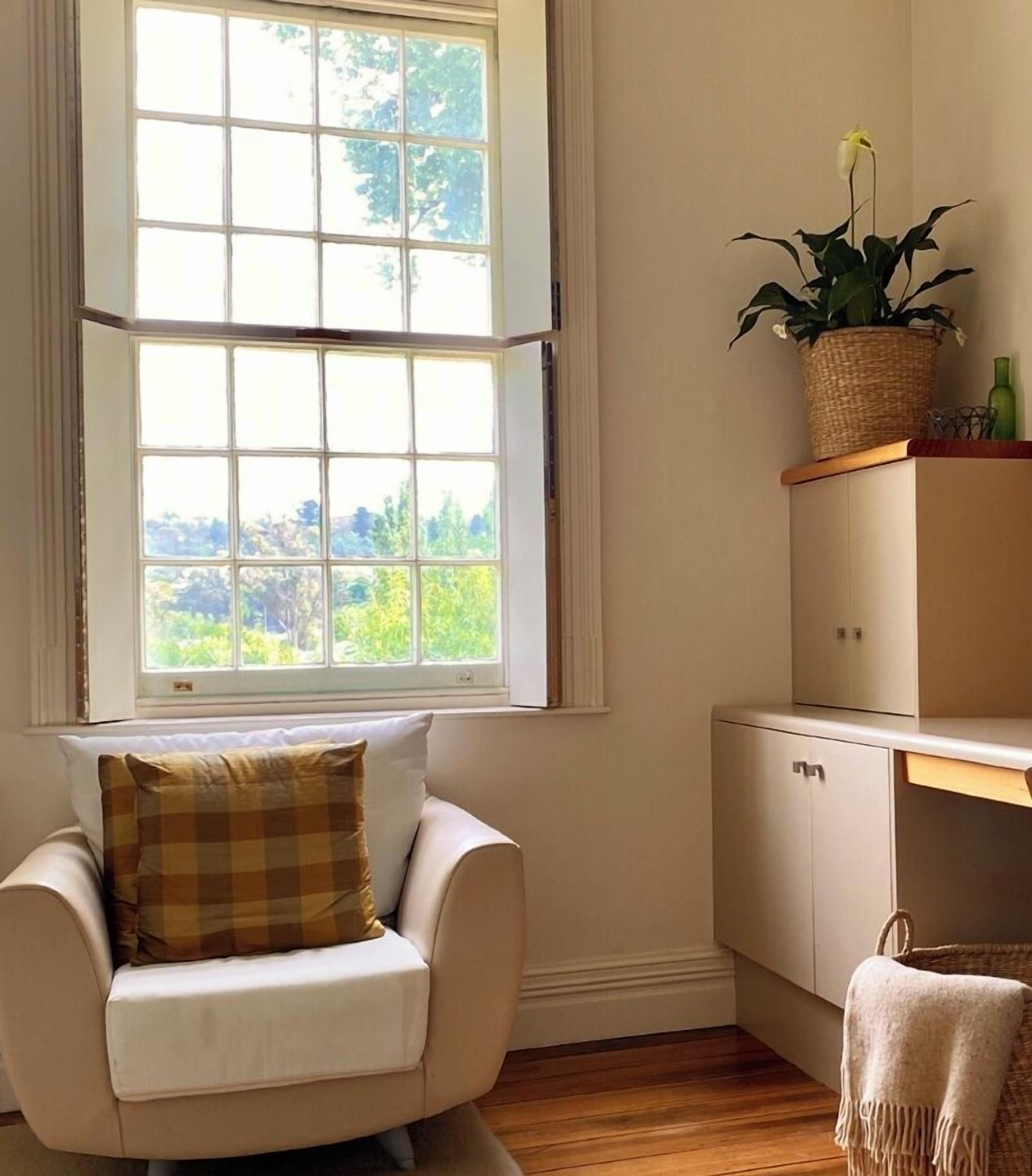 A corner room with a white armchair, a small cabinet, a window with a view of greenery, and a potted plant on a shelf.