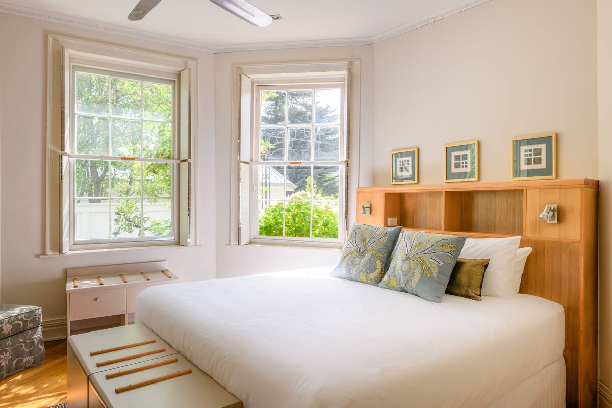 Een slaapkamer met een groot bed, twee ramen, een houten hoofdbord en een nachtkastje.