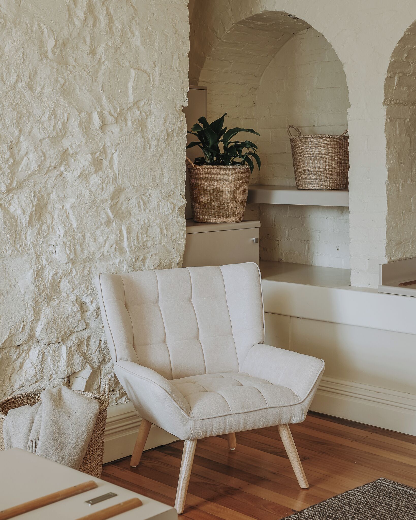 A room with a white upholstered armchair, a wooden floor, a white textured wall, and a wicker basket on a shelf.