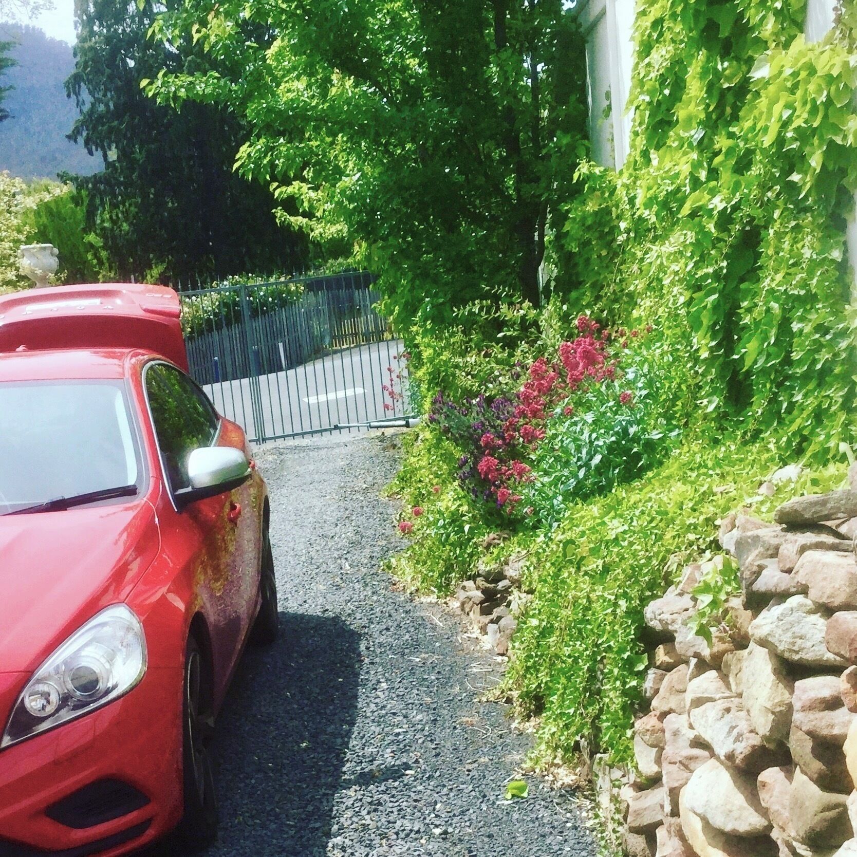 A red car parked on a narrow road with a stone wall on one side and a metal fence on the other.