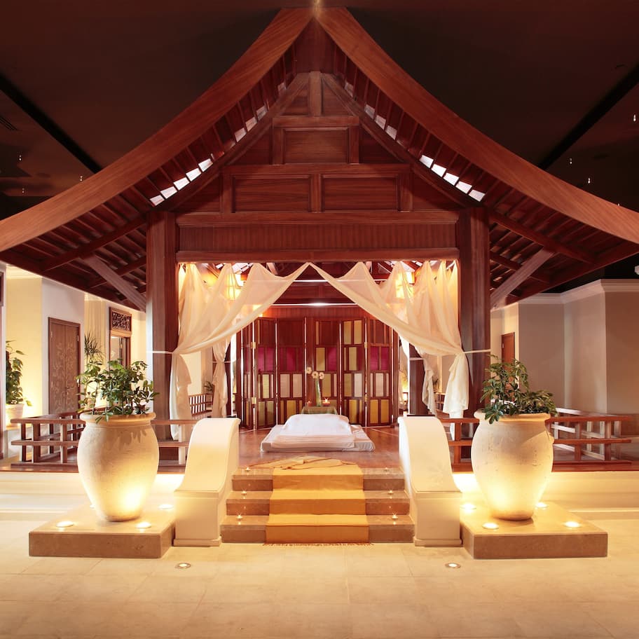 A traditional wooden structure with a bed at the center, flanked by large potted plants and a staircase leading up.