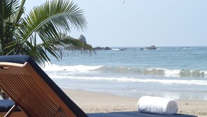 A beach with wooden lounge chairs and a rolled towel, palm trees, and a rocky outcrop in the water. - Pacífica Resort Ixtapa All-Inclusive (Ixtapa)