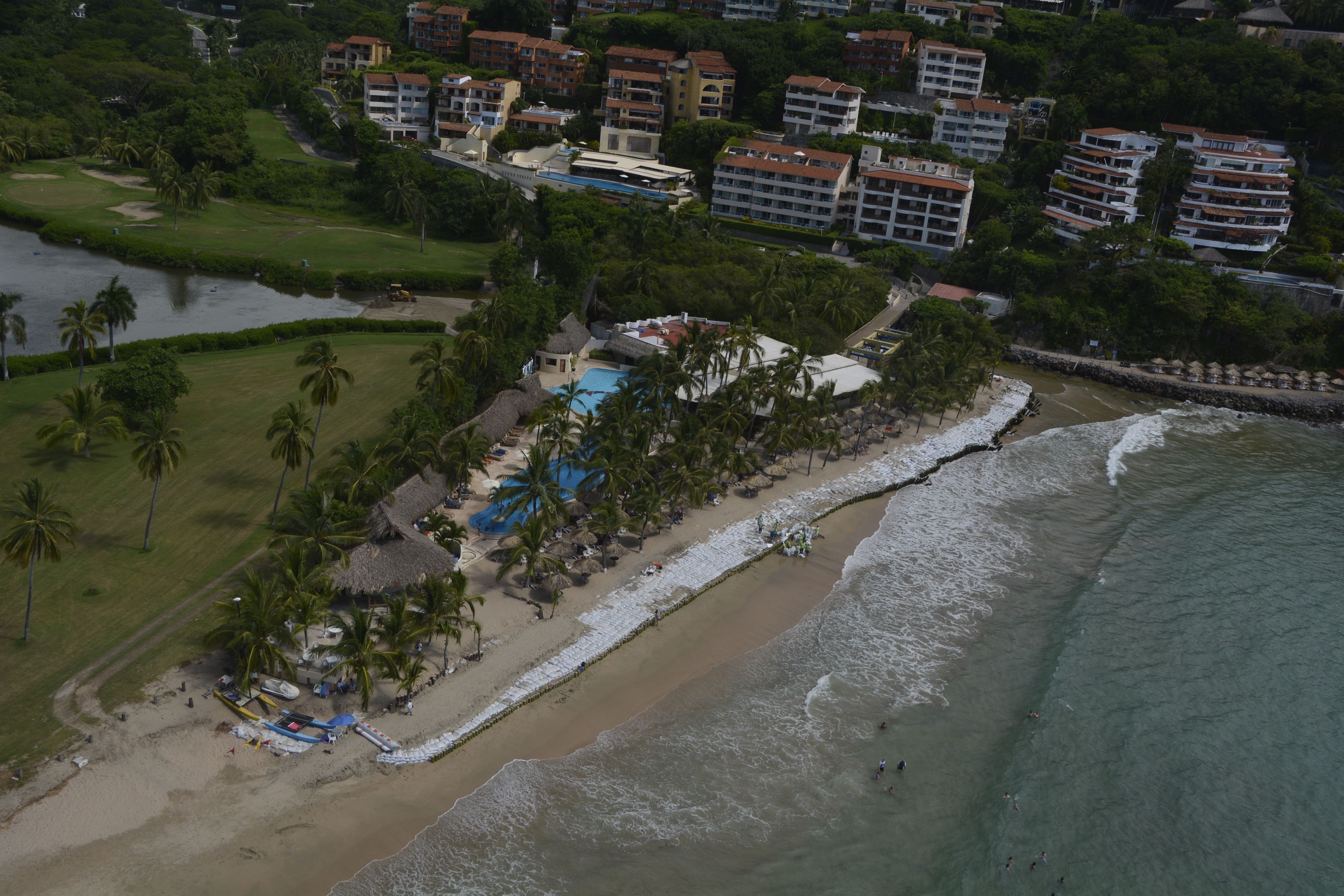 An aerial view of a coastal resort with multiple hotel buildings, a beach, and a pool area surrounded by palm trees.