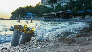 A beach with a metal bucket containing bottles, a glass bottle, and a metal cup. There is a building with multiple balconies and a beachside restaurant with umbrellas. - Hotel Pappas (Loutraki-Agioi Theodoroi)