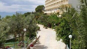 A hotel with a long, straight pathway leading to a pool area, surrounded by lush greenery and a view of the sea. - Hotel Pappas (Loutraki-Agioi Theodoroi)