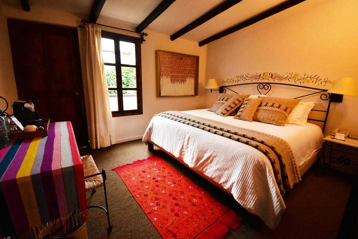 Um quarto com uma cama, uma mesa com toalha listrada, um tapete vermelho e uma janela com cortinas.