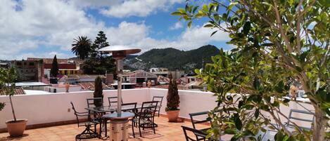 Terraza en la azotea con mesa y sillas, plantas en macetas y vistas a un paisaje urbano y montañas.