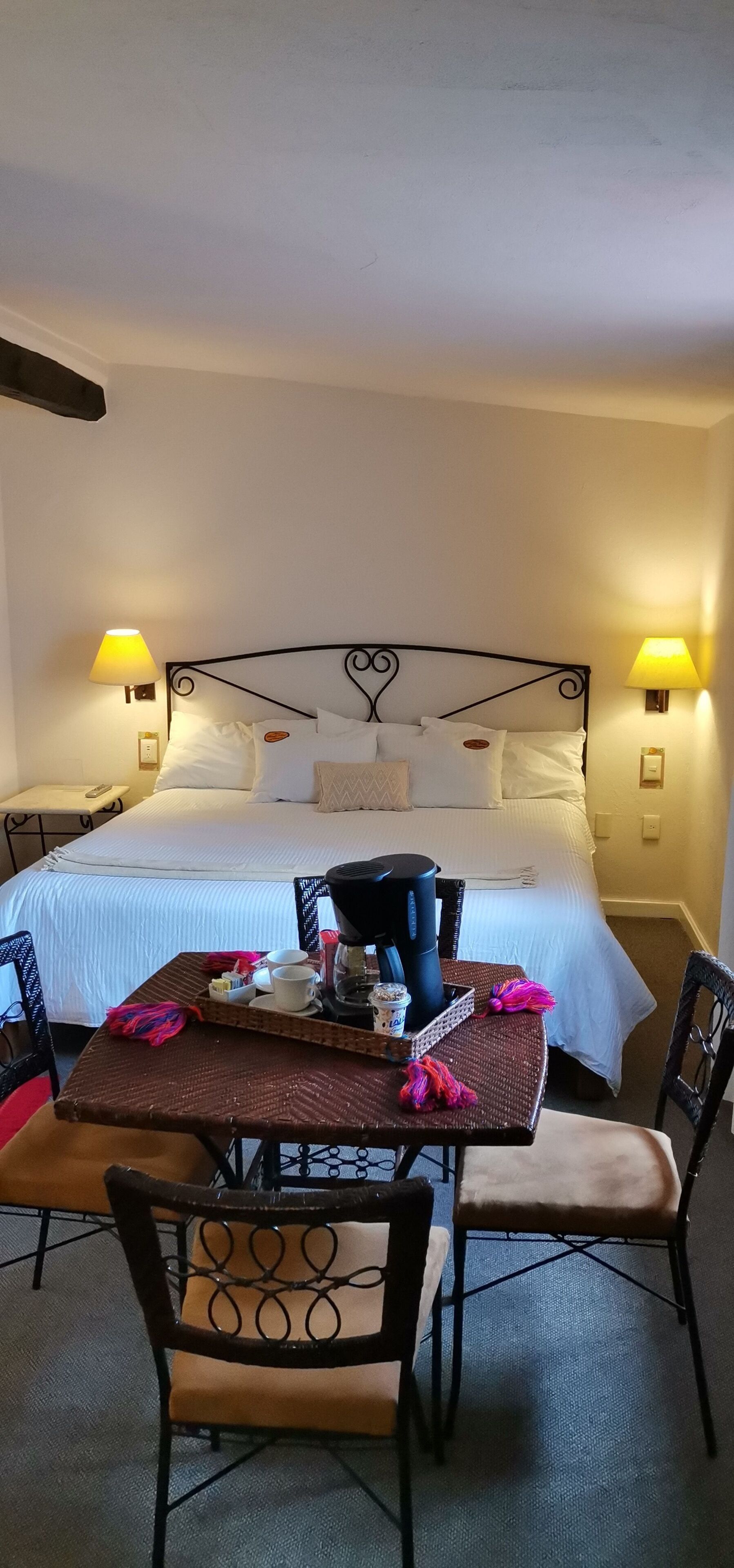 Een hotelkamer met een bed, nachtlampjes, een tafel met een koffiezetapparaat, kopjes en een dienblad.