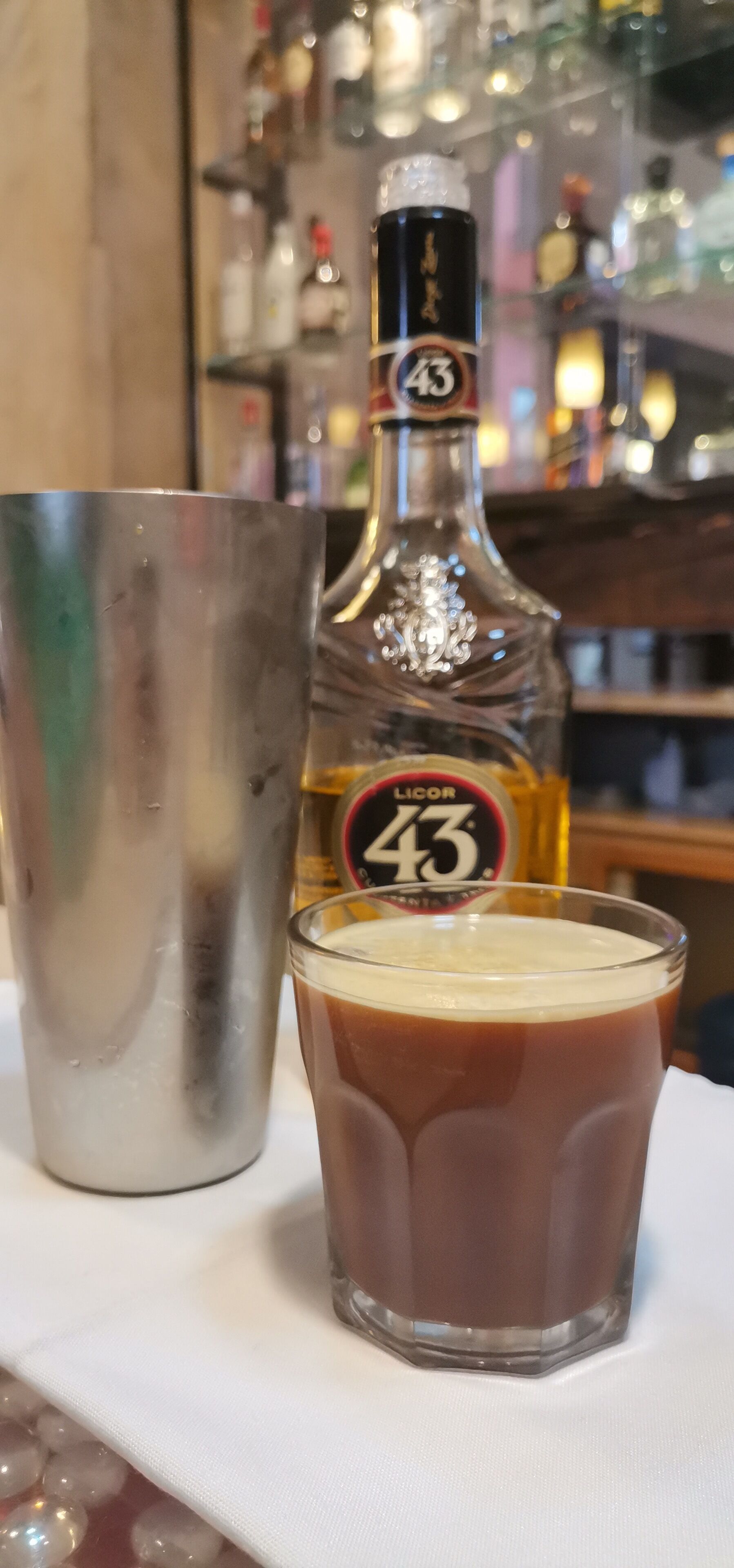 Et glas med gyldent væske, en metalkværn og en flaske med etiket i en bar.