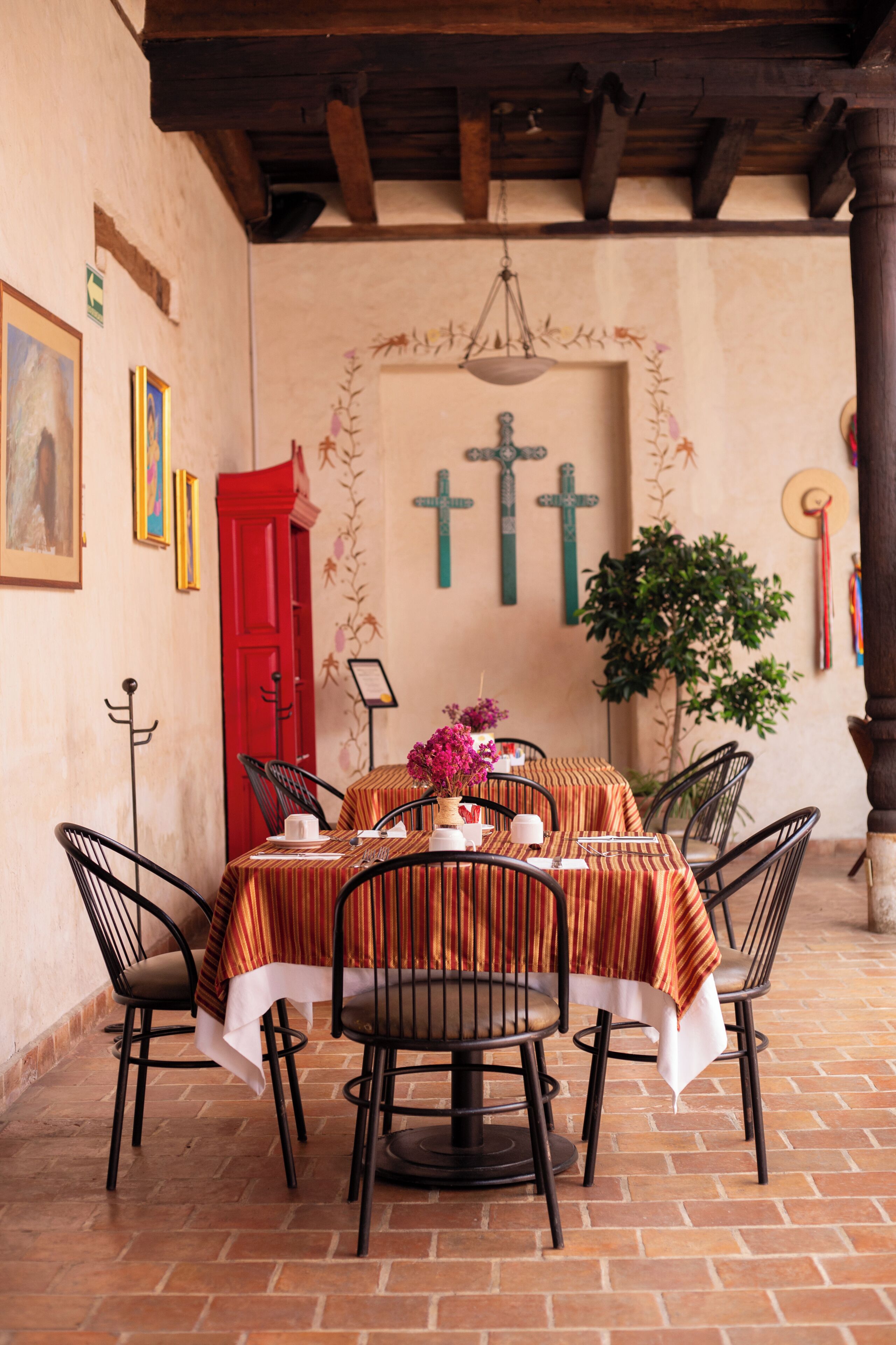 Uma sala de jantar com uma mesa posta para quatro pessoas, rodeada de cadeiras, uma porta vermelha e uma parede com uma cruz e plantas.