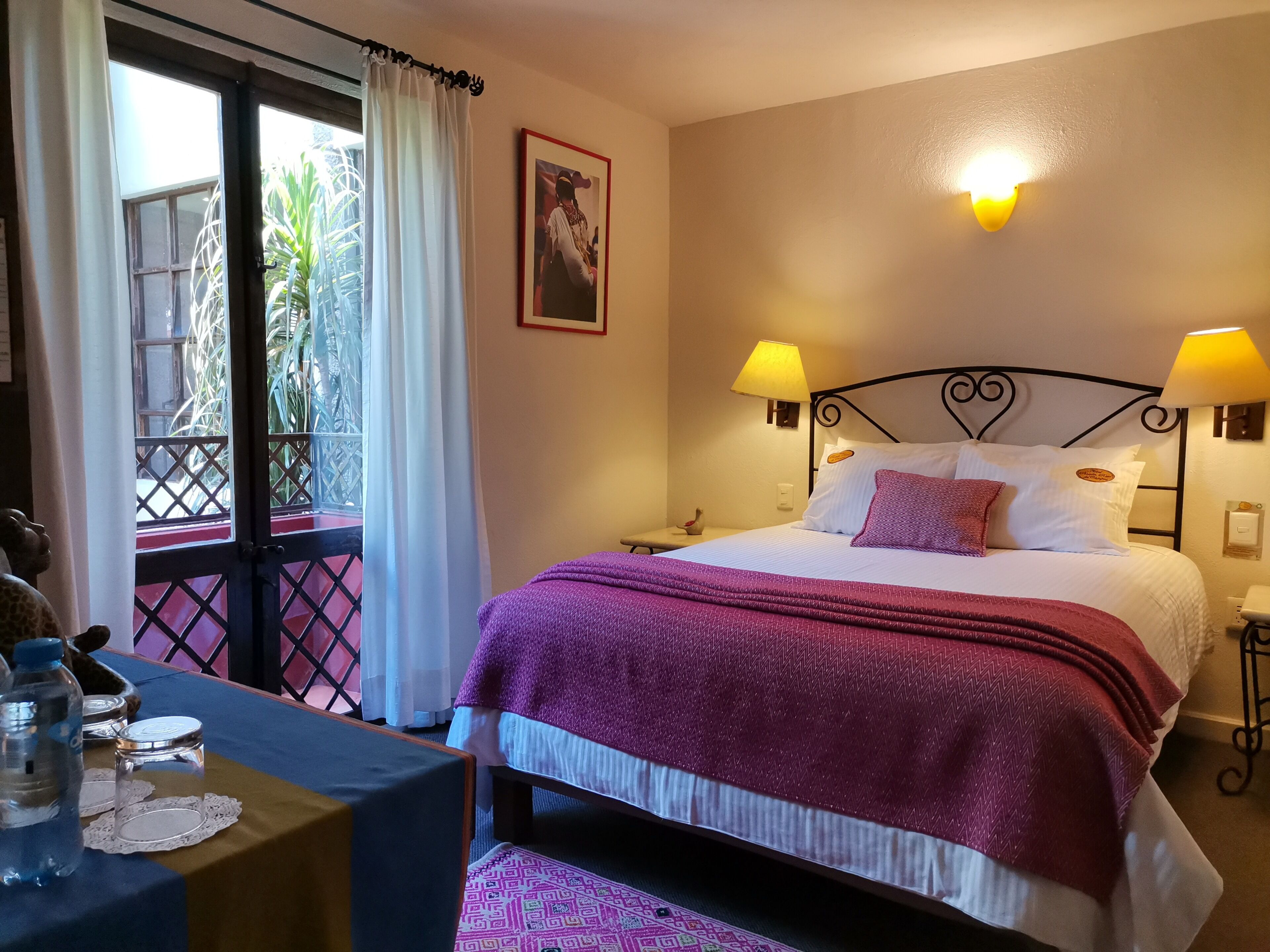 A hotel room with a bed, two bedside lamps, a nightstand, a desk with a water bottle, and a view of the outside through a window with curtains.