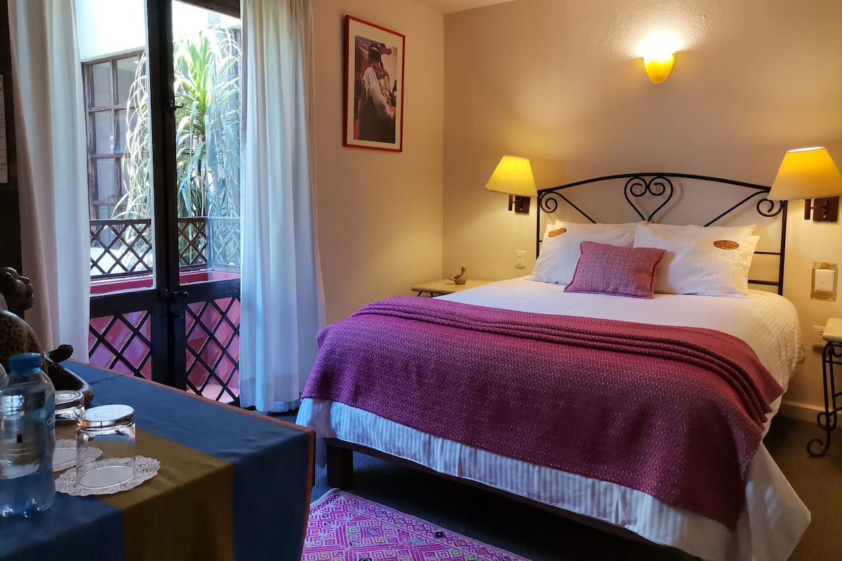 A hotel room with a bed, two bedside lamps, a nightstand, a desk with a water bottle, and a view of the outside through a window with curtains.