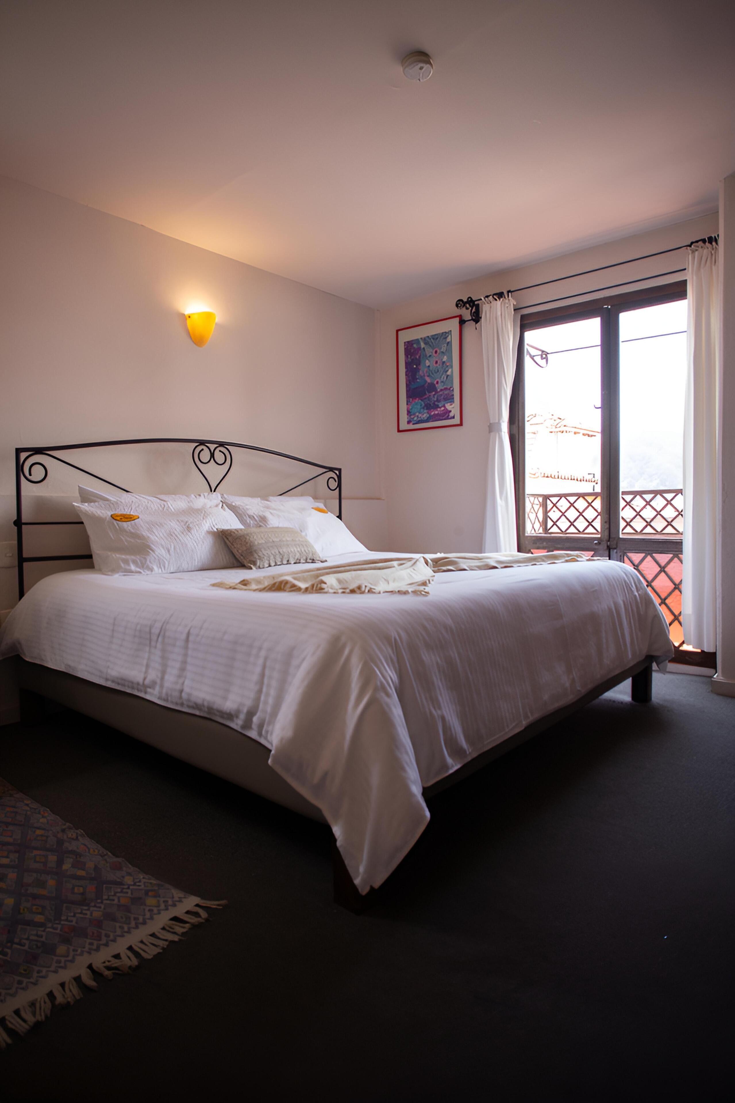 A bedroom with a bed, a wall-mounted light, a window with curtains, and a balcony view.