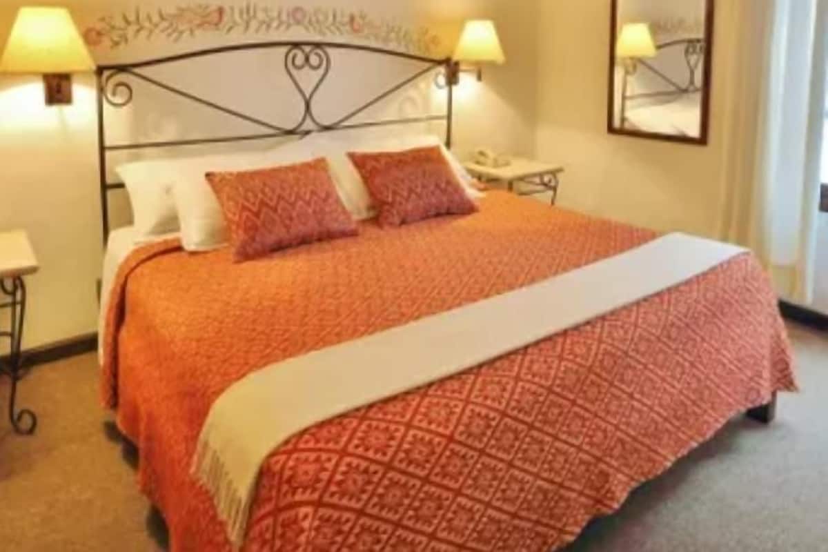 Quarto de hotel com cama, duas luminárias de cabeceira, mesa de cabeceira, espelho e janela com cortinas.