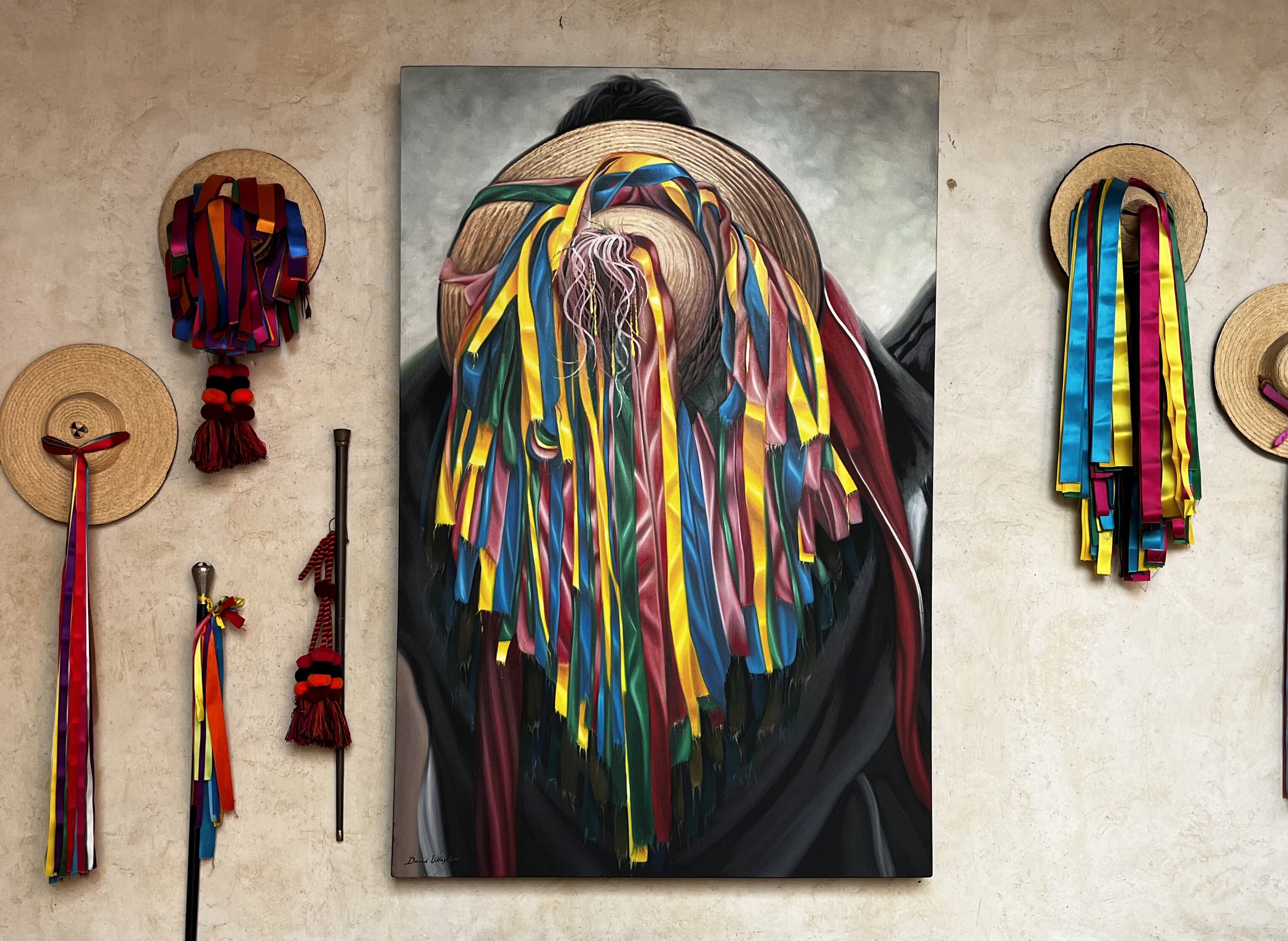 Una pintura que muestra a una persona con una llamativa decoración en la cabeza y un atuendo negro, rodeada de cintas y sombreros de colores sobre un muro con textura.