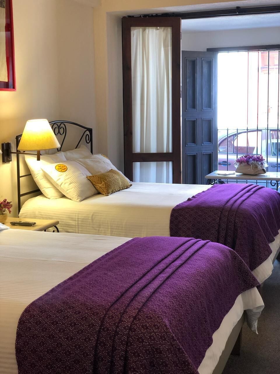 Une chambre d’hôtel avec deux lits, une table de chevet, une lampe, une fenêtre avec des rideaux et une vue sur la rue.
