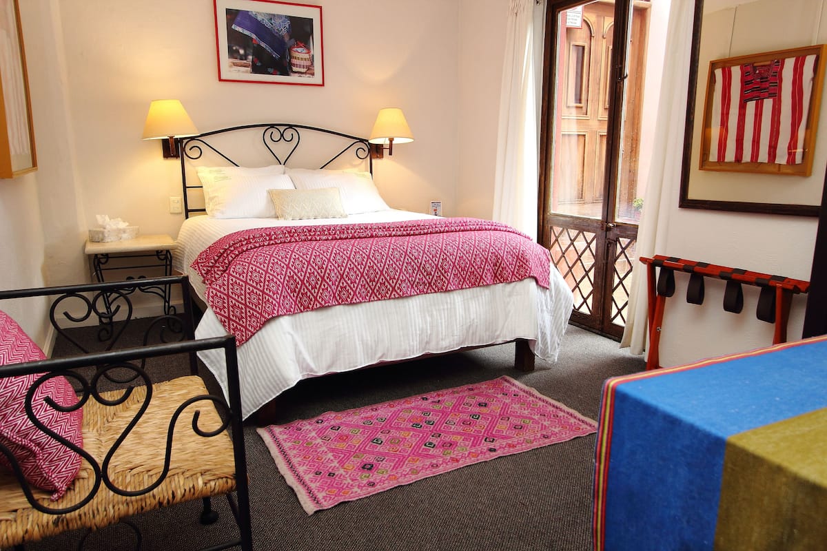 Quarto de hotel com cama, duas luminárias de cabeceira, uma mesinha, uma cadeira e uma janela com cortinas.