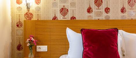 Una cama bien hecha con un cojín rojo y un florero de flores rosas sobre una mesita de noche de madera.