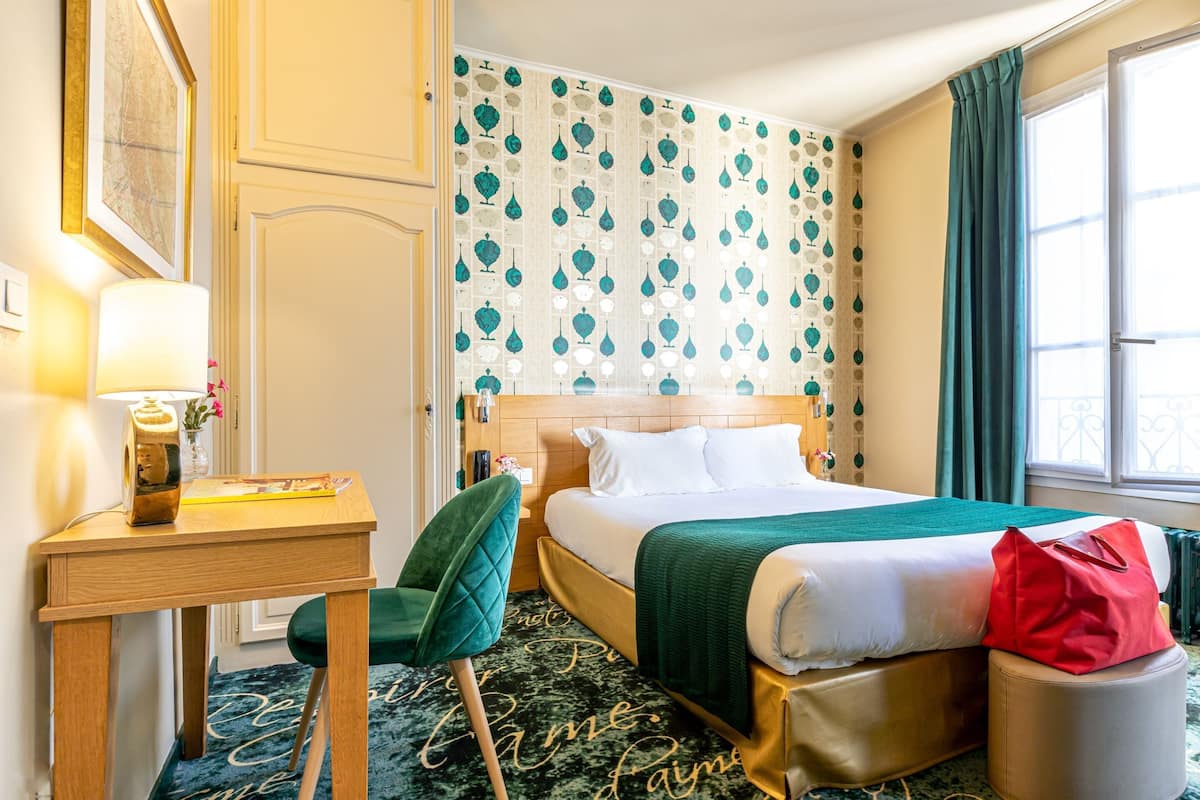 Habitación de hotel con cama, escritorio, silla, bolso rojo y ventana con cortinas.