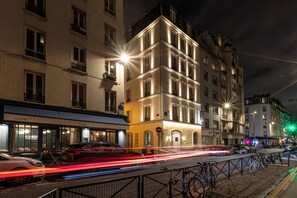 A nighttime street scene with illuminated buildings, a restaurant named 'Roche', and bicycles parked along the sidewalk.