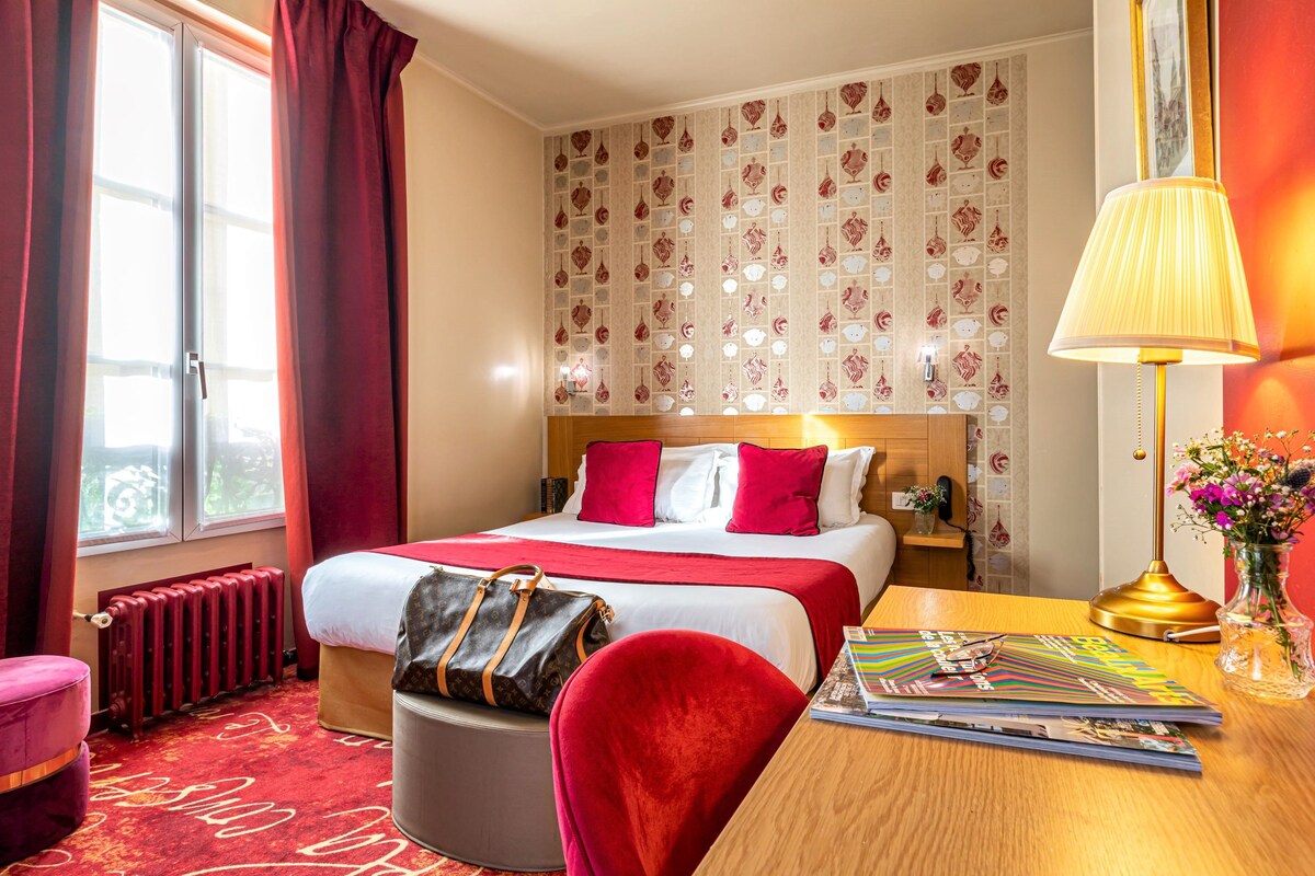 Habitación de hotel con cama, escritorio, silla, lámpara, bolso y radiador.