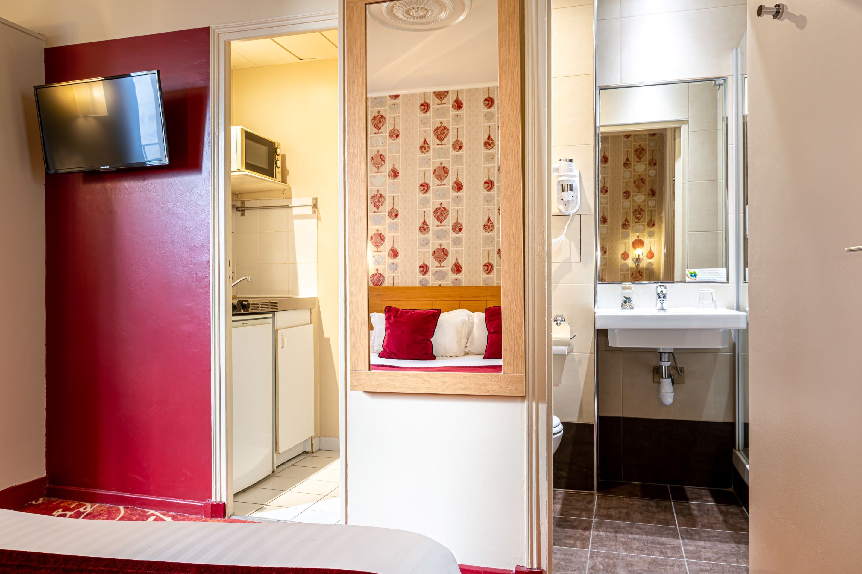 Hotelkamer met een rode accentmuur, een aan de muur bevestigde televisie, een bed met rode en witte beddengoed, een kleine badkamer met spiegel en wastafel, en uitzicht op de aangrenzende kamer.