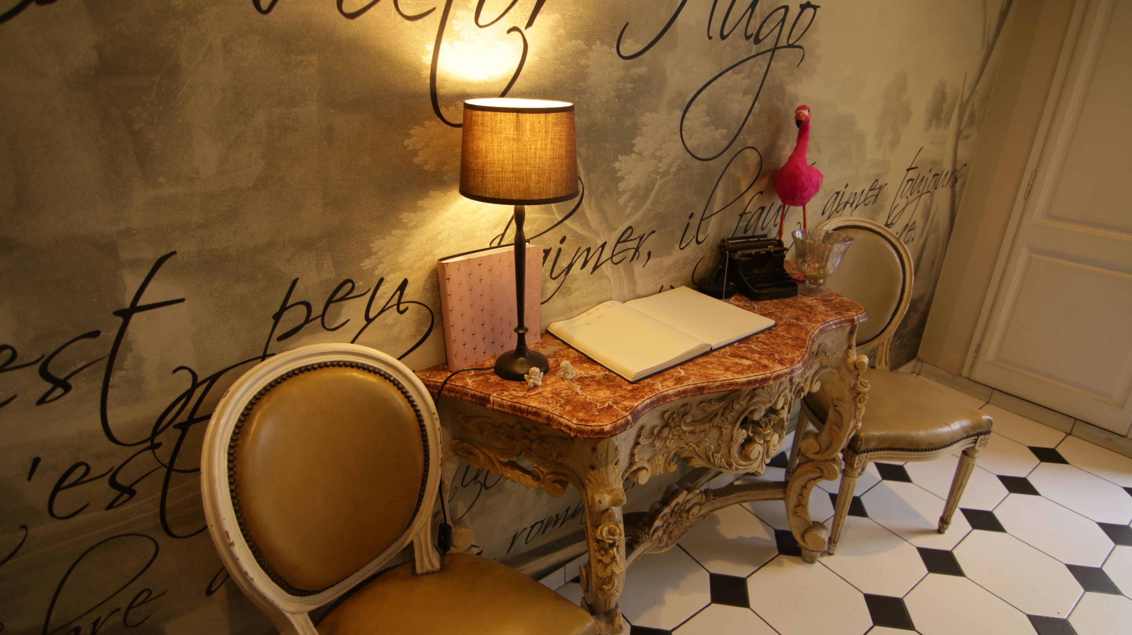 A room with a marble table, two chairs, a lamp, a typewriter, and a decorative flamingo.