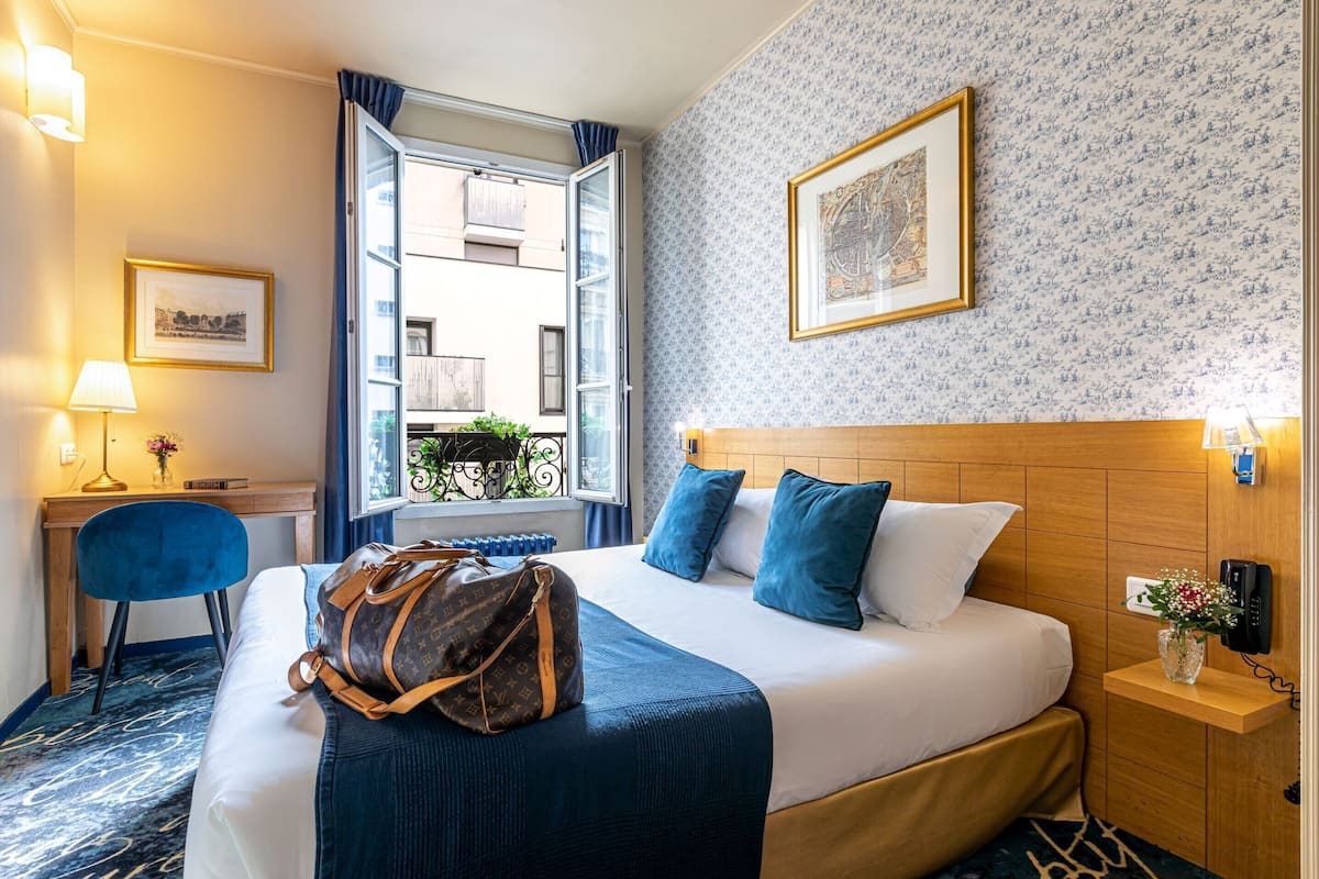 Habitación de hotel con cama, escritorio, silla, ventana y pared con un cuadro enmarcado.