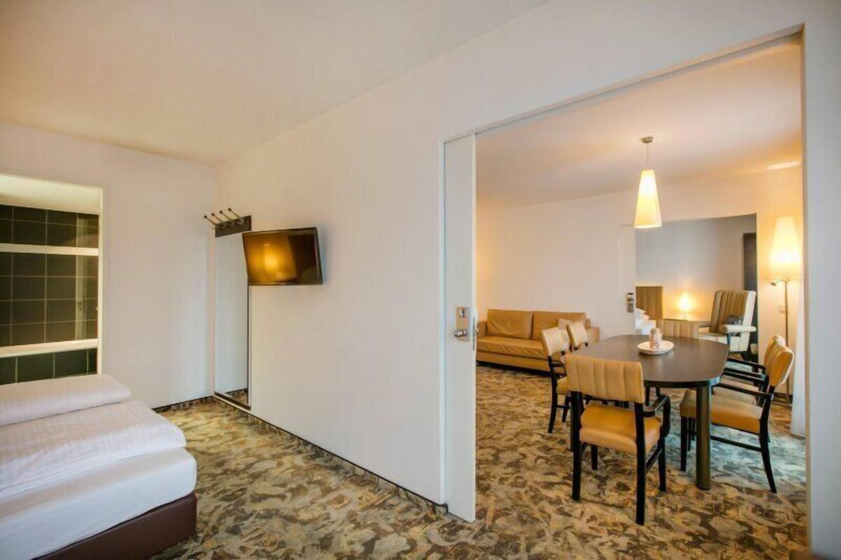Quarto de hotel com cama, televisão na parede, área de jantar com mesa e cadeiras, e um sofá.