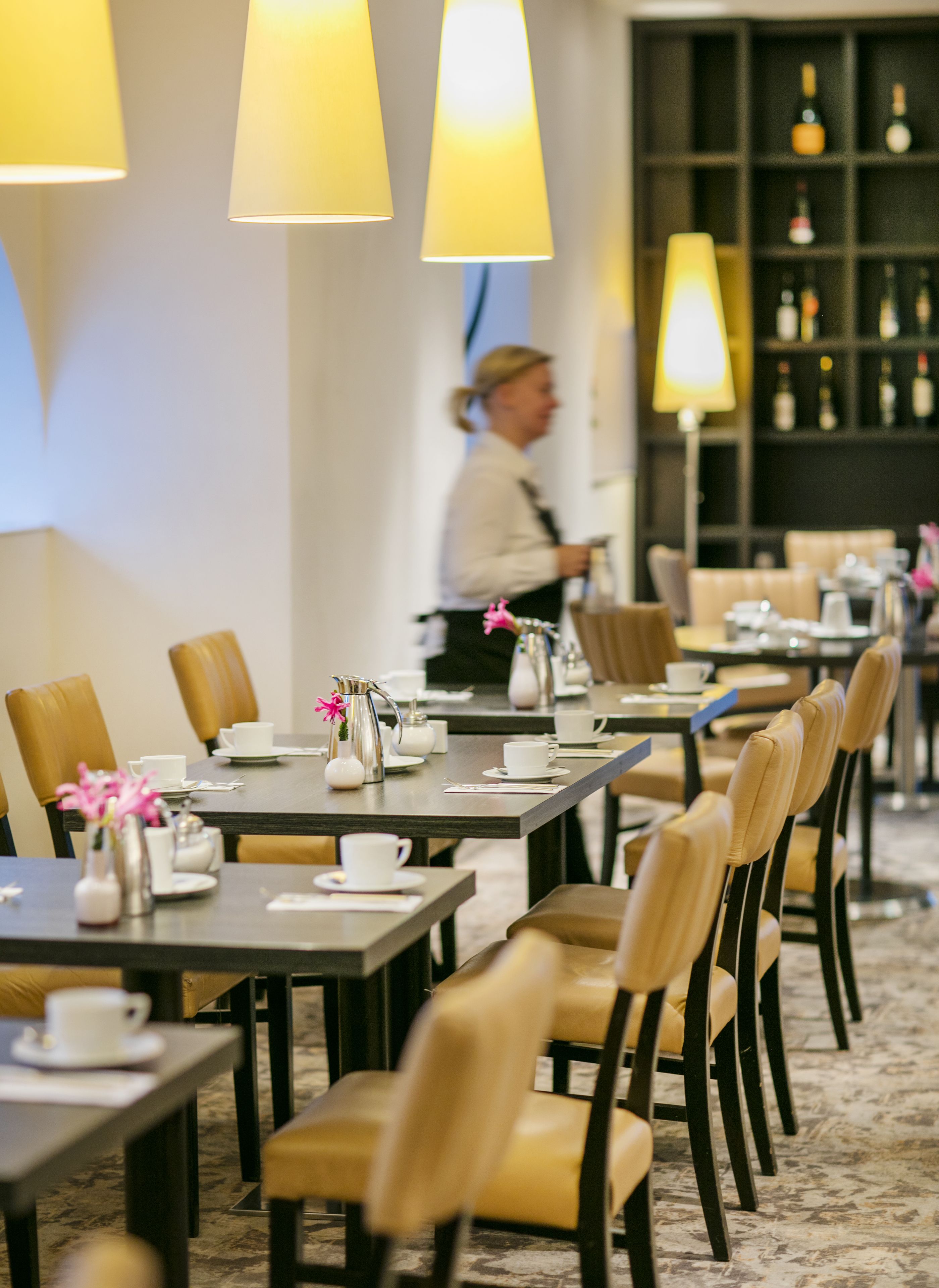 Un comedor de hotel con mesas preparadas para el servicio, una zona de bar con botellas y una persona al fondo.