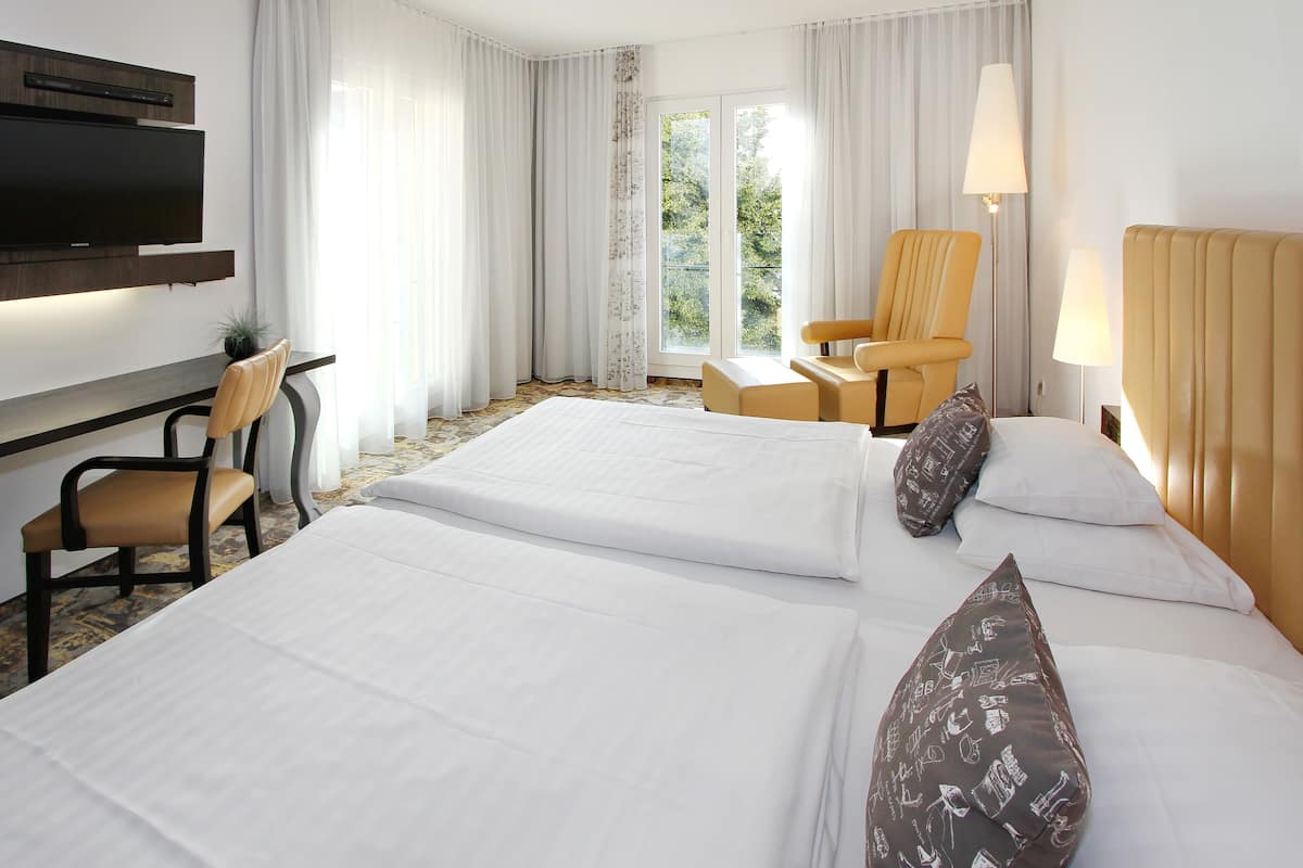 Quarto de hotel com duas camas, uma escrivaninha, uma cadeira, uma televisão e uma janela com cortinas.