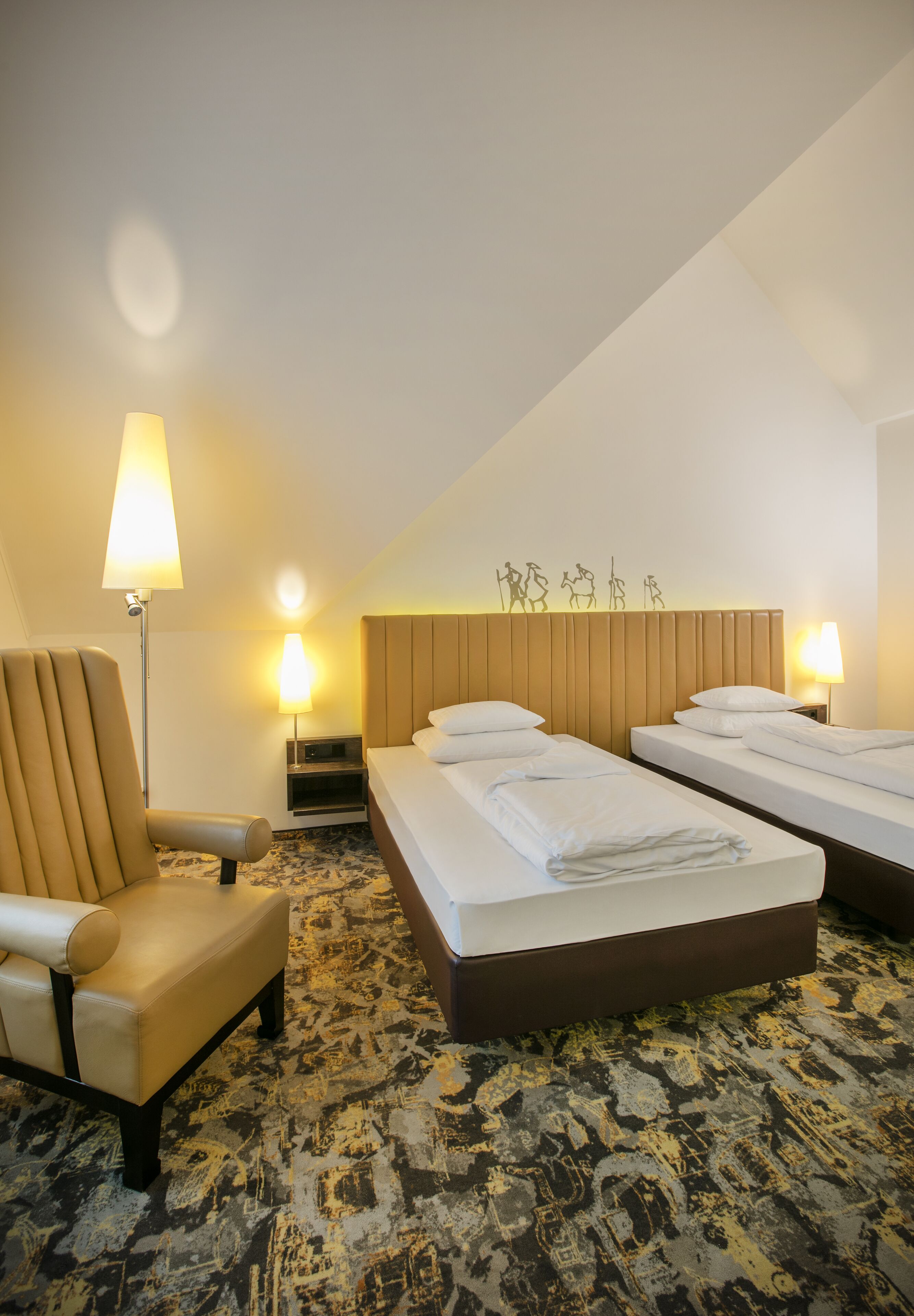 A hotel room with two beds, a chair, a lamp, and a patterned carpet.