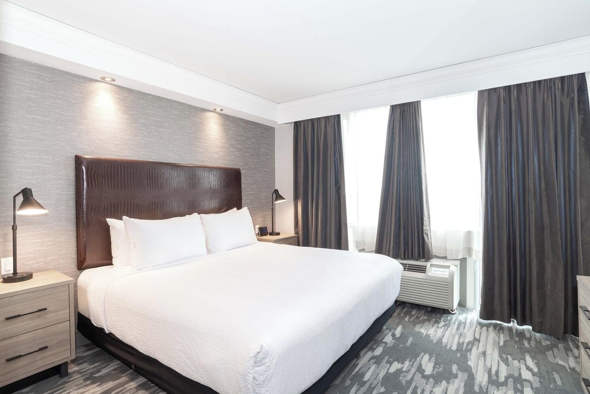 A hotel room with a large bed, two bedside tables, a wall-mounted air conditioning unit, and a window with curtains.