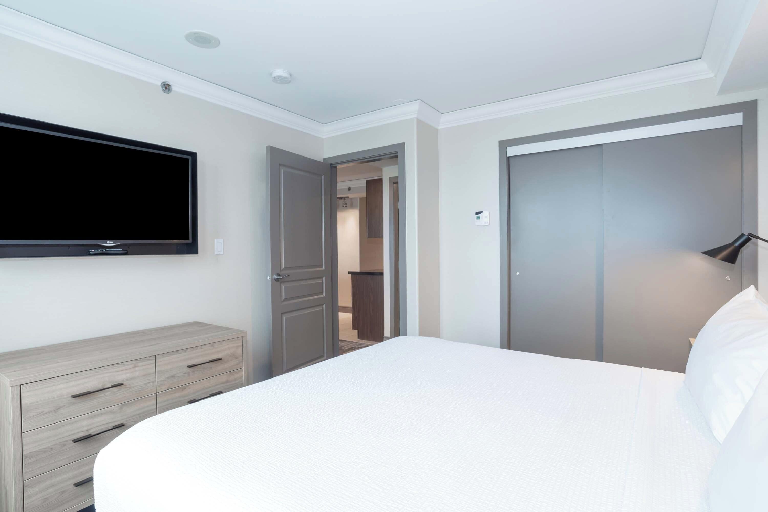 A hotel room with a bed, a flat-screen TV mounted on the wall, a wooden dresser, and a closet.