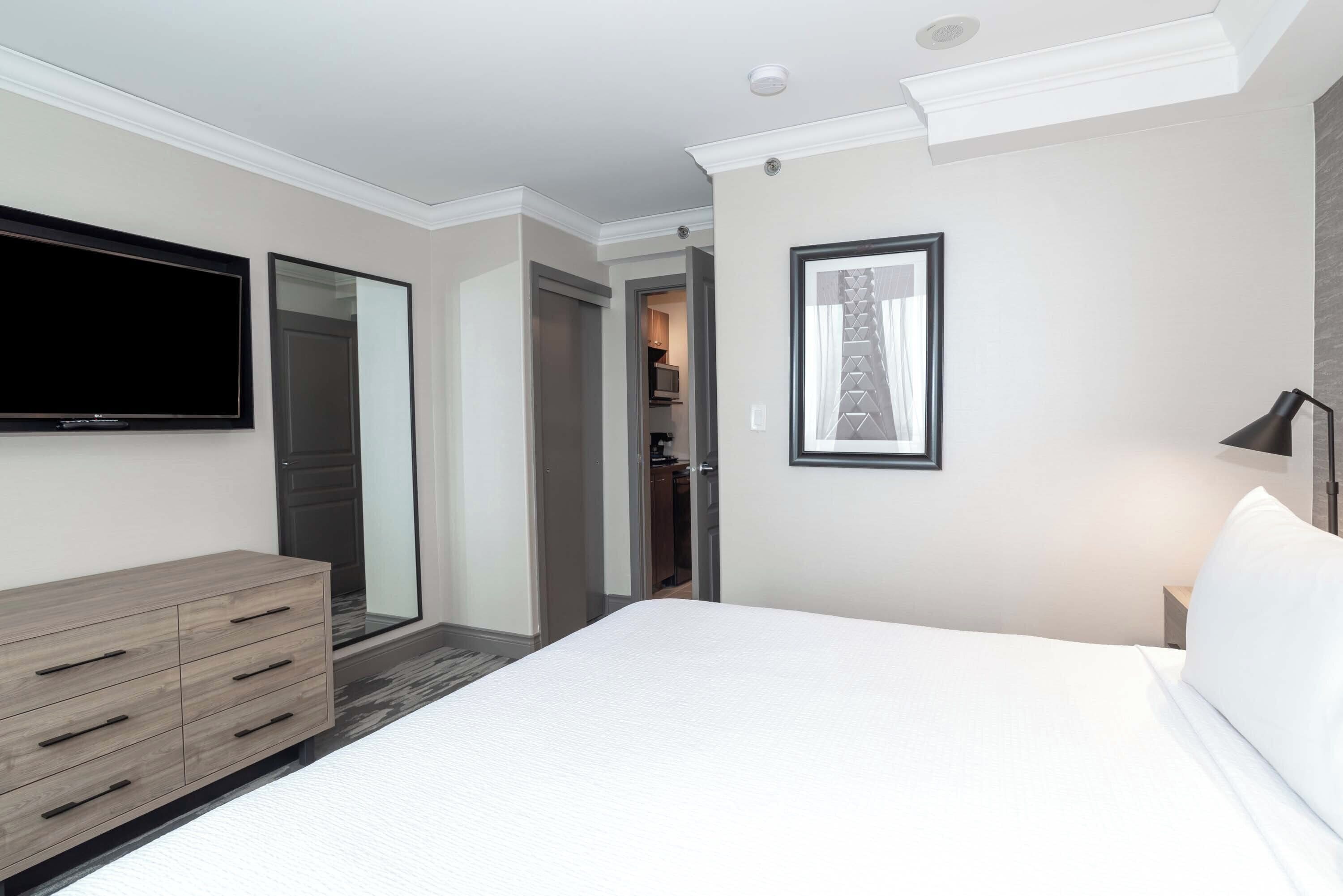 A modern hotel room with a large bed, a flat-screen TV mounted on the wall, a wooden dresser, and a framed picture.